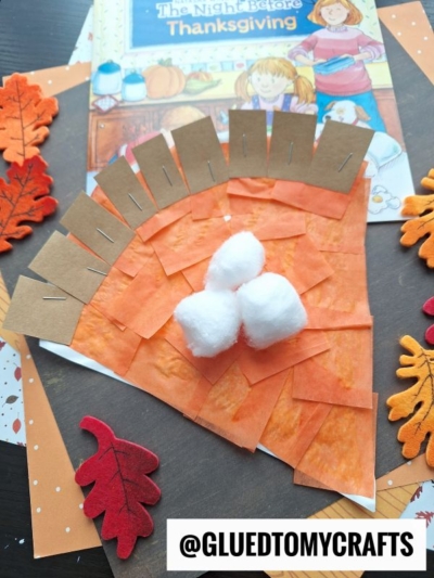 A child’s Thanksgiving pumpkin pie paper plate craft made with orange and brown paper, cotton balls, and felt leaves sits on display. A book titled The Night Before Thanksgiving is in the background. The image is watermarked @GLUEDTOMYCRAFTS.