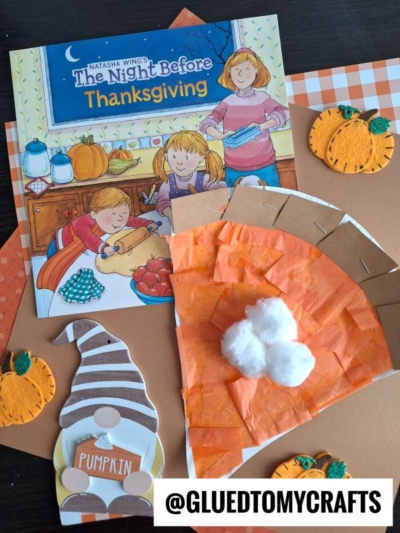 A children’s book titled The Night Before Thanksgiving is surrounded by autumn-themed crafts, including a pumpkin pie paper plate craft, felt pumpkins, and a pie slice made from orange tissue paper and cotton balls, all on a dark table.