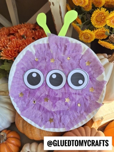 A Paper Plate Monster craft featuring a purple alien with three big eyes, green antennae, and gold star stickers, surrounded by orange flowers and pumpkins. The @GLUEDTOMYCRAFTS username is displayed at the bottom.