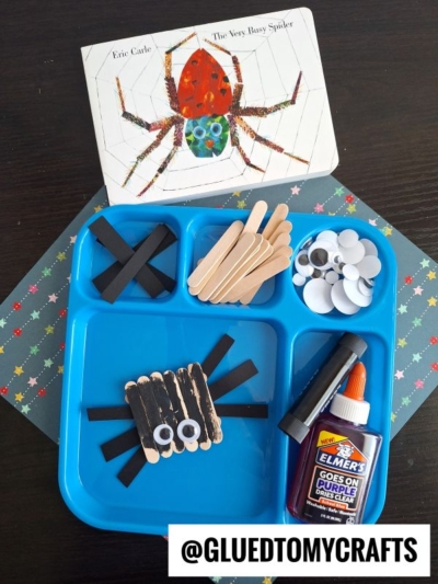A blue tray holds black paper strips, craft sticks, googly eyes, and glue for a craft stick spider activity. The Very Busy Spider book sits at the top, and a finished popsicle stick spider craft is displayed on the tray.