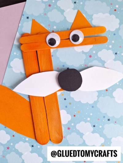 A fox crafted from orange popsicle sticks, white and black paper, and googly eyes is placed on a blue, cloud-patterned background. This F is for craft stick fox project features the text @GLUEDTOMYCRAFTS at the bottom.