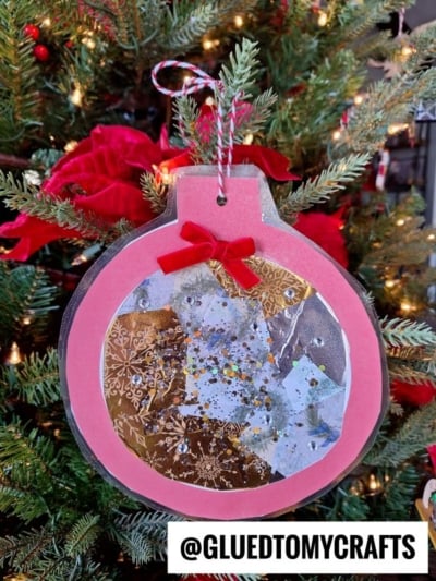 A handmade stained glass ornament with a pink border, sparkly paper pieces, and a red bow hangs on a decorated tree. The text @GLUEDTOMYCRAFTS is at the bottom.
