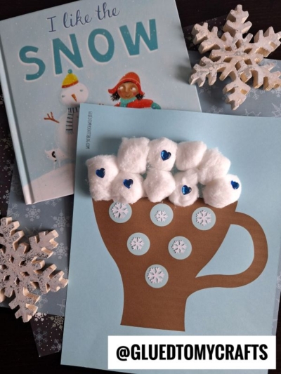 A children’s book called I Like the Snow is next to a winter craft of a paper hot cocoa mug on blue paper, decorated with cotton ball marshmallows and snowflake stickers. Wooden snowflake ornaments are nearby.