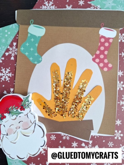 A handprint Christmas fireplace craft features two stockings, a sparkling gold-glitter fire, and a Santa cutout. A @GLUEDTOMYCRAFTS label accents the bottom, with snowflake-patterned paper as the festive background.