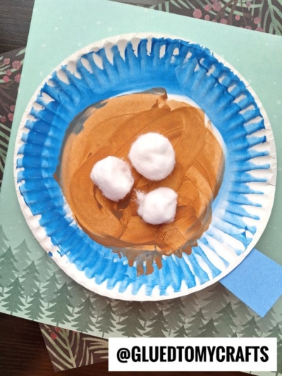 A paper plate painted blue and brown holds three cotton balls, resembling a nest with eggs—much like a cozy paper plate hot cocoa mug project. The plate sits on a green paper with tree designs, while a printed label reads @GLUEDTOMYCRAFTS.
