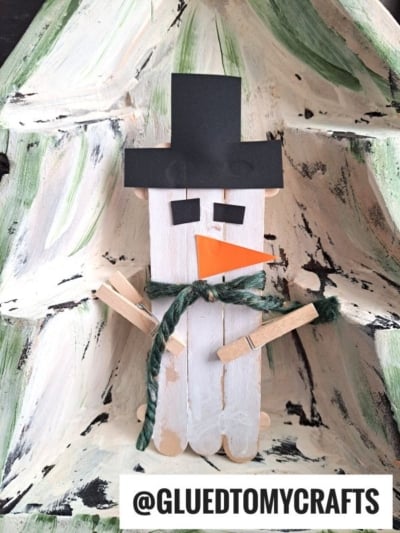 A Popsicle Stick Snowman craft featuring a black paper hat, orange paper nose, green yarn scarf, and clothespin arms, displayed on a rustic painted wooden background. @GLUEDTOMYCRAFTS text is shown below.