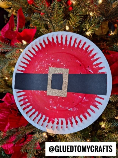 A festive holiday craft featuring a paper plate Santa belly painted red with a black strip and gold glitter buckle, displayed in front of a decorated Christmas tree. Text at bottom reads “@GLUEDTOMYCRAFTS.”.
