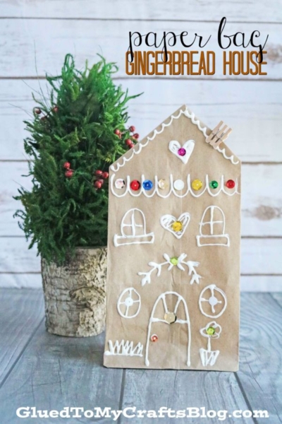A small faux Christmas tree in a rustic pot sits beside a charming gingerbread house paper bag craft, decorated with white icing designs and colorful sequins.