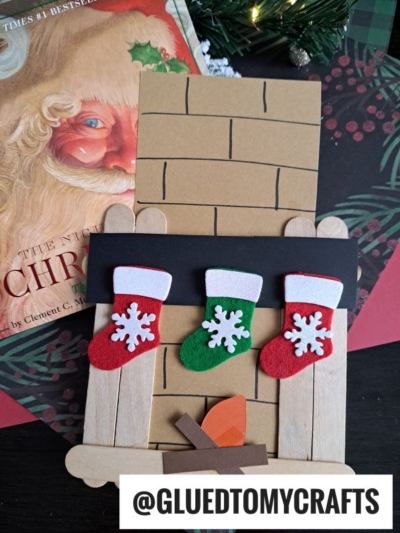 A handmade popsicle stick fireplace craft, adorned with three felt stockings and a small paper fire, sits next to a Christmas book featuring Santa on the cover. The handle @GLUEDTOMYCRAFTS is displayed.