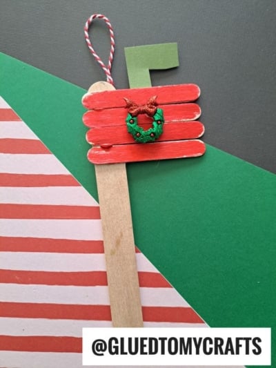 A handmade popsicle stick mailbox ornament shaped like a red mailbox, featuring a small green wreath and striped string. The handle reads @GLUEDTOMYCRAFTS. Background includes festive red, green, and striped paper.