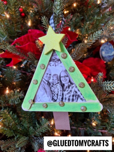 Handmade popsicle stick Christmas tree frame ornament in a triangle shape, featuring a smiling family photo, gold glitter dots, and a yellow star. Perfect for hanging among red decorations! Text: @GLUEDTOMYCRAFTS.