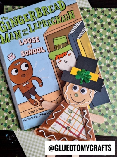 A handmade popsicle stick gingerbread man leprechaun craft with paper and googly eyes sits beside the book The Gingerbread Man and the Leprechaun Loose at School. The Instagram handle @GLUEDTOMYCRAFTS is displayed at the bottom.
