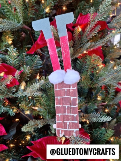 A popsicle stick Santa stuck in a chimney ornament, featuring two red legs in gray boots and white pom-pom trim, hangs cheerfully on a decorated Christmas tree.