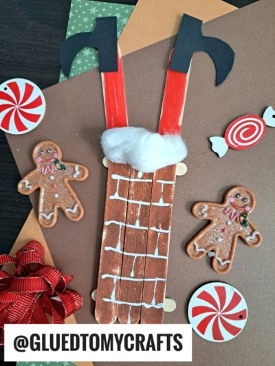 A festive craft project featuring a popsicle stick Santa stuck in a chimney with cotton snow, surrounded by paper gingerbread men, candy decorations, and a red bow—all set against a cozy brown background.