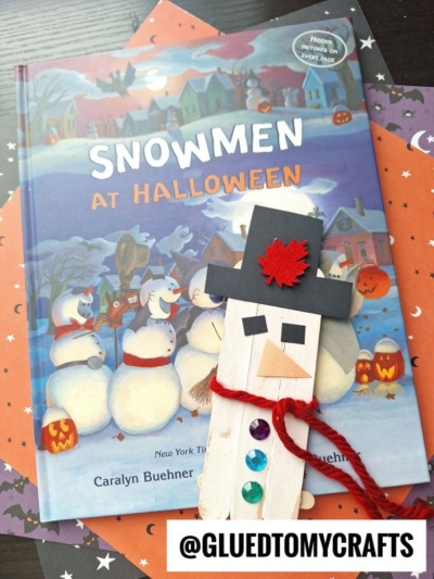 A children’s book titled Snowmen at Halloween lies on star-patterned paper, with a craft stick snowman bookmark—made from a popsicle stick, decorated with gems, a red scarf, and a black hat. @GLUEDTOMYCRAFTS is written below.