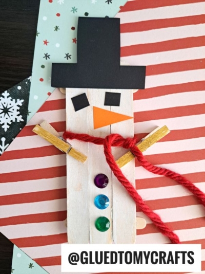 A Popsicle Stick Snowman featuring a red yarn scarf, clothespin arms, a black paper hat, and button decorations, is placed on patterned paper. Text reads @GLUEDTOMYCRAFTS.