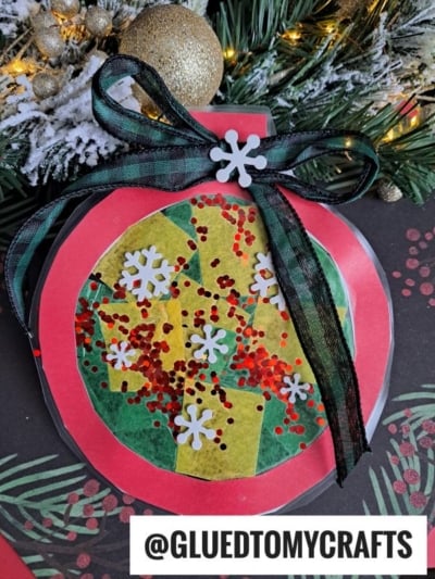 A handmade Christmas ornament craft inspired by stained glass window ornaments, made from red and green paper with glitter, tissue paper, white snowflake cutouts, and topped with a black and green plaid ribbon bow. Pine branches and gold ornaments decorate the background.
