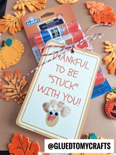 A tag reading “thankful to be Stuck With You!” with googly eyes and a painted thumbprint turkey is tied to Elmer’s glue sticks, surrounded by felt pumpkins and autumn leaves. Text @GLUEDTOMYCRAFTS at the bottom.