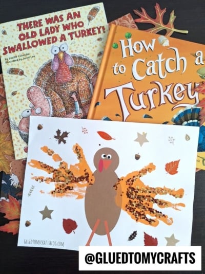 A children's craft shows a handprint turkey keepsake made from two orange painted handprints as wings, with a brown oval body, red legs, googly eyes, an orange beak, and glittered handprint turkey feathers on white paper.
