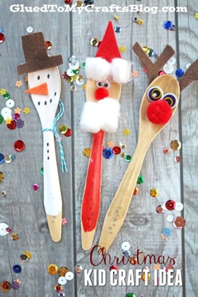 Three wooden spoons decorated as a snowman, Santa, and a wooden spoon reindeer with felt, pom-poms, and googly eyes are surrounded by colorful sequins on a wooden surface. Text reads Christmas Kid Craft Idea.