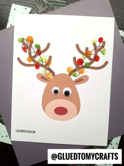 A thumbprint reindeer craft made from paper, featuring large googly eyes and antlers decorated with colorful buttons, swirly black lines, and thumbprint Christmas lights on a white background. The Instagram handle @GLUEDTOMYCRAFTS is at the bottom.