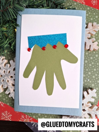 A handmade toddler handprint Christmas card features a green handprint cutout resembling reindeer, with a blue glitter strip and red embellishments on top, surrounded by festive paper, snowflakes, and pine branches.