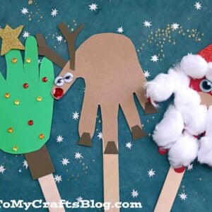 Three Christmas puppets made from paper and craft sticks: a handprint reindeer, a handprint Christmas tree with decorations, and a handprint Santa with a cotton ball beard, all set on a blue background with stars.