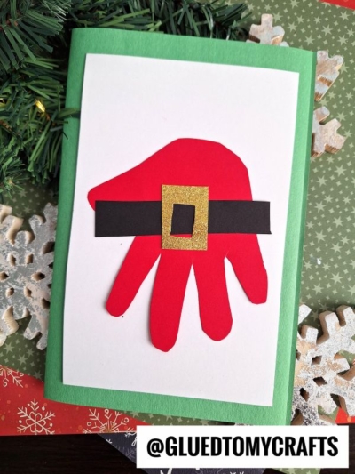 A green handprint Christmas card decorated with a red paper handprint, designed to look like Santa’s suit with a black belt and gold buckle, placed on a festive background with snowflakes and pine branches.