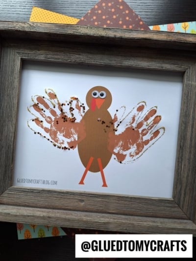 A framed turkey craft featuring a brown paper turkey with orange legs and beak, googly eyes, and handprint turkey feathers dusted in gold glitter. Colorful patterned paper shows behind the frame. @GLUEDTOMYCRAFTS tag is at bottom.