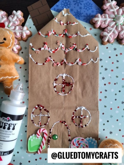A brown paper bag gingerbread house is decorated with puffy paint, surrounded by felt gingerbread, candy canes, cookies, and a puffy paint bottle. Snowflake-patterned paper is used as the background to complete this charming gingerbread house paper bag craft scene.