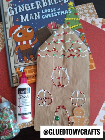 A gingerbread house paper bag craft, decorated with white paint and glitter, sits on a table with The Gingerbread Man Loose at Christmas book, craft supplies, and festive patterned paper. @GLUEDTOMYCRAFTS is written at the bottom.
