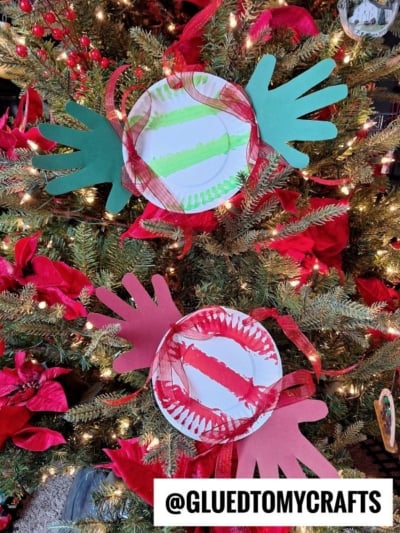 Two paper plate Christmas ornaments, inspired by handprint peppermint candy, with painted stripes and cut-out hand shapes, decorated with red ribbon, hang on a tree with red flowers and lights. @GLUEDTOMYCRAFTS is written at the bottom.