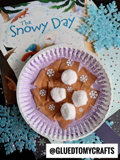 A paper plate hot cocoa mug craft inspired by The Snowy Day features brown paper squares, white cotton balls, and snowflake confetti, surrounded by blue glitter snowflakes and the book in the background. Text: @GLUEDTOMYCRAFTS.