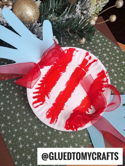 A festive craft featuring a white paper plate peppermint candy with red paint stripes, two red ribbon bows, and blue handprint cutouts, placed on a green star-patterned background near holiday decorations.