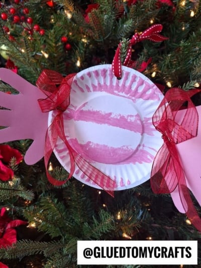A paper plate peppermint candy craft featuring red stripes, pink handprint cutouts on each side, and red ribbon bows, hangs on a Christmas tree. Text at the bottom reads @GLUEDTOMYCRAFTS.