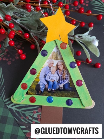 A handmade popsicle stick Christmas tree frame ornament, painted green and adorned with gems and a yellow star, beautifully surrounds a family photo. Red berries and greenery are in the background. @GLUEDTOMYCRAFTS is at the bottom.