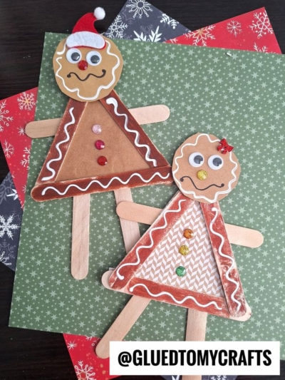 Two craft stick gingerbread men made from popsicle sticks, paper, and googly eyes, decorated with sequins and hats, are placed on festive patterned paper. @GLUEDTOMYCRAFTS is written at the bottom.