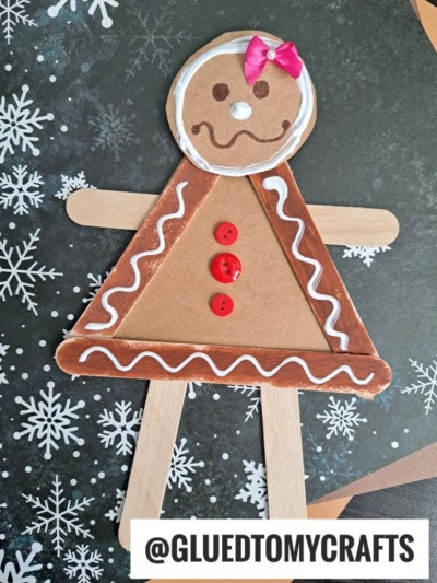 A craft stick gingerbread girl made from popsicle sticks, with red buttons, white icing designs, and a pink bow, displayed on a snowflake background. Text: @GLUEDTOMYCRAFTS.
