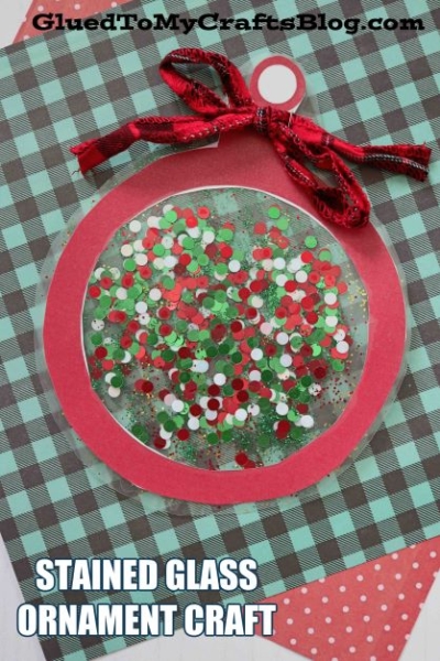 A handmade stained glass christmas ornament crafted from paper, clear plastic, and festive confetti in red, green, and white. Finished with a red ribbon on top, this lovely piece is perfect for those who love stained glass ornaments. Displayed on a checkered background.