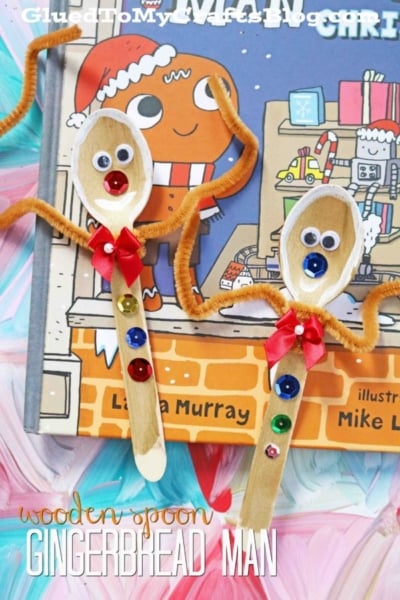 Two wooden spoons decorated as gingerbread men with googly eyes, red bows, pipe cleaner arms, and colorful buttons are placed on a children’s Christmas book featuring a cartoon gingerbread character. These charming creations are perfect for holiday crafting alongside other favorites like wooden spoon reindeer or a cheerful wooden spoon snowman.