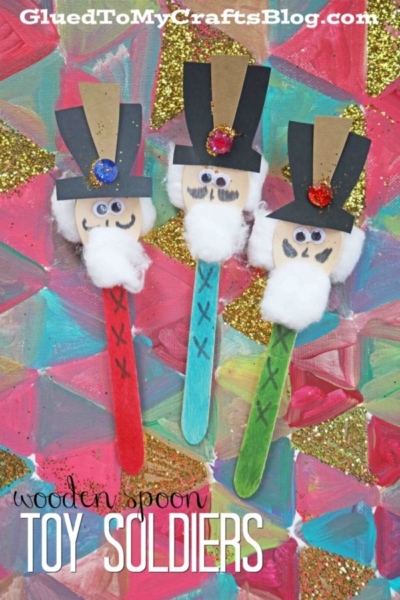 Three colorful wooden spoons decorated as toy soldiers with hats, googly eyes, felt beards, and jewels are placed on a glittery, abstract pink and gold background. These charming figures pair perfectly with other festive crafts like a wooden spoon snowman or wooden spoon christmas friends. Text reads: wooden spoon TOY SOLDIERS.