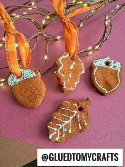 Four acorn-shaped applesauce Christmas ornaments with orange ribbons and white icing details rest on a pink surface. A branch with string lights and the handle @GLUEDTOMYCRAFTS appears at the bottom.