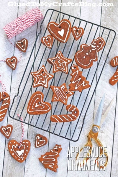 Assorted heart, star, candy cane, tree, and snowman-shaped applesauce Christmas ornaments with white icing rest on a cooling rack, alongside twine and scissors on a rustic white surface. Text reads APPLESAUCE ORNAMENTS.