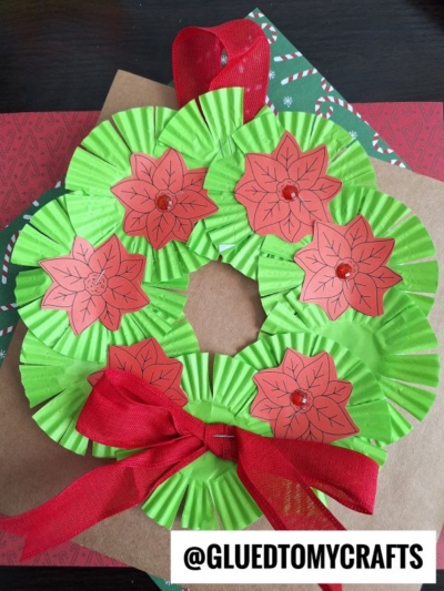 A handmade cupcake liner Christmas wreath features green paper liners, red poinsettias, a ribbon bow, and is displayed on festive paper. The handle @GLUEDTOMYCRAFTS appears at the bottom—perfect for your next Christmas wreath craft!.