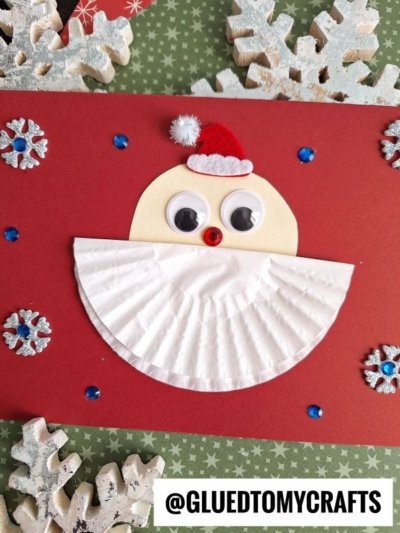 A handmade Santa card features a red background, semicircle face with googly eyes, small red pom-pom nose, and cotton-topped hat. A folded cupcake liner Santa beard completes the look, with snowflake decorations scattered around.