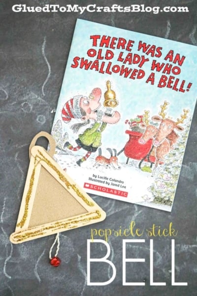 A childrenās book titled There Was an Old Lady Who Swallowed a Bell! and a handmade popsicle stick Christmas bell ornament with a red bead lay on a gray background. Text at the bottom reads, popsicle stick BELL.