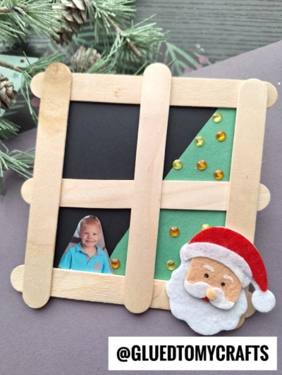 A handmade Christmas photo frame made of craft sticks features a felt Santa, gold sequins, a child’s photo, and @GluedToMyCrafts text. A popsicle stick Christmas tree in window design adds festive charm, while green pine branches fill the background.