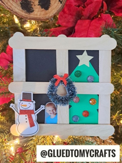 A handmade Christmas frame ornament with craft sticks features a snowman cutout, a child’s photo, a wreath, and a decorated green tree—plus a popsicle stick Christmas tree in the window—hanging on a festive Christmas tree. Text reads @GLUEDTOMYCRAFTS.