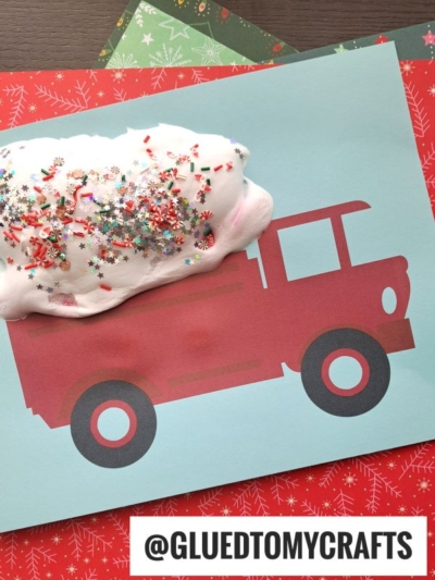 A red fire truck silhouette on blue paper with white foam, puffy paint snow, and colorful glitter on top, simulating snow. The background includes festive patterned papers. Text at the bottom reads @GLUEDTOMYCRAFTS.