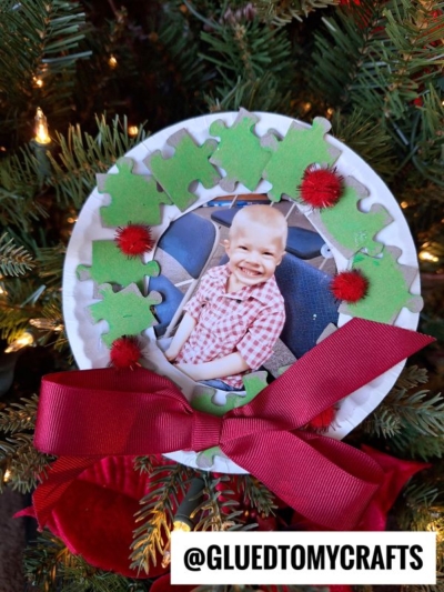 A handmade Puzzle Piece Christmas Wreath photo ornament hangs on a tree, featuring a child’s smiling photo framed by green puzzle pieces, red pom-poms, and a festive red bow. The username @GLUEDTOMYCRAFTS appears at the bottom.